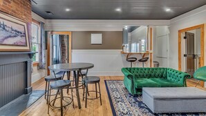 A modern living room with a fireplace, a green sofa, a round table with stools, and a patterned rug. - Charming 2BR w/ Porch • Historic Stay Near Forsyth Park (Savannah)