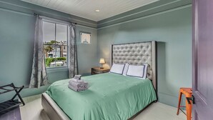 A bedroom with a large bed, a nightstand with a lamp, a window with curtains, and a view of buildings outside. - Charming 2BR w/ Porch • Historic Stay Near Forsyth Park (Savannah)