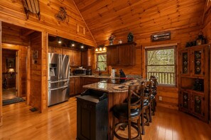A cabin-style kitchen with wooden walls, a central island with bar stools, stainless steel appliances, and a window with a view. - Mountain Escape w/ Hot Tub & Game Room (Sevierville)