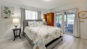 Un dormitorio con cama, dos mesitas de noche, una mesilla, una ventana con cortinas, una puerta que da a un espacio exterior y una planta en la pared.