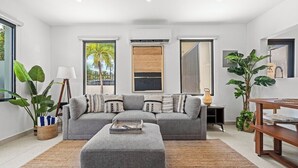 Ein modernes Wohnzimmer mit einem grauen Sofa, einem Couchtisch und einem großen Fenster mit Blick auf Palmen.