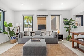 A modern living room with a grey sofa, a coffee table, and a large window with a view of palm trees. - Villa Bali: Private Pool | 2 Min Walk to Beach (San Juan)