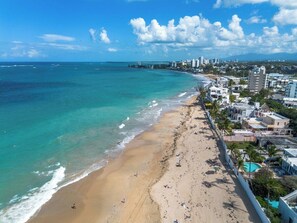 A beach with a clear view of the ocean and a cityscape in the background. - Villa Bali: Private Pool | 2 Min Walk to Beach (San Juan)