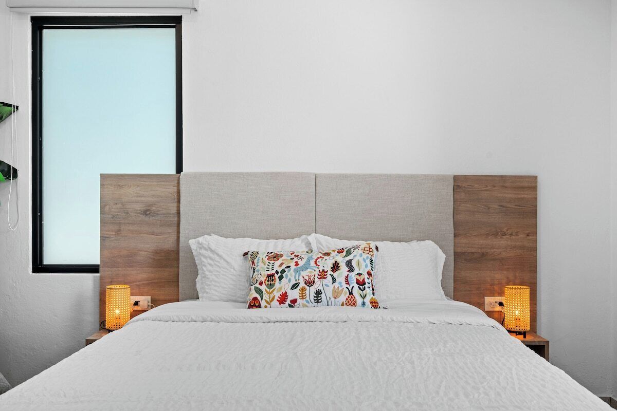 A neatly made bed with a white duvet and two pillows, a wooden headboard, bedside lamps, and a window with blinds.