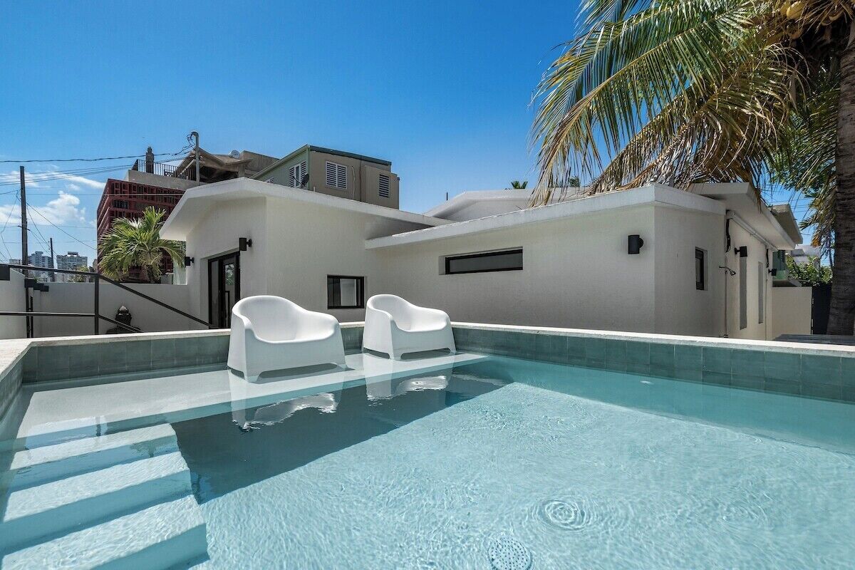A modern building with a swimming pool, two white lounge chairs, and a palm tree.