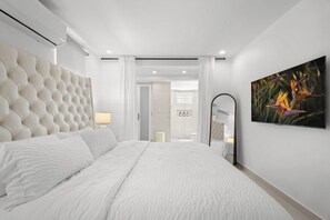 A modern bedroom with a tufted headboard, a large bed with white linens, a flat-screen TV mounted on the wall, and a bathroom visible through an open door. - Villa Tropical: Private Pool | 2 Min Walk to Beach (San Juan)