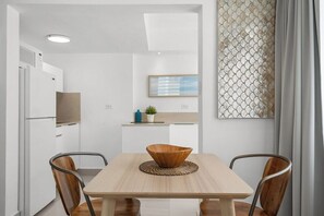 A modern kitchen with white cabinetry, a wooden dining table with a woven placemat, and a view of the living area through an open door. - Villa Tropical: Private Pool | 2 Min Walk to Beach (San Juan)