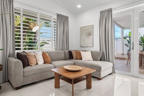 A modern living room with a grey sectional sofa, a wooden coffee table, and a large window with blinds. - Villa Tropical: Private Pool | 2 Min Walk to Beach (San Juan)