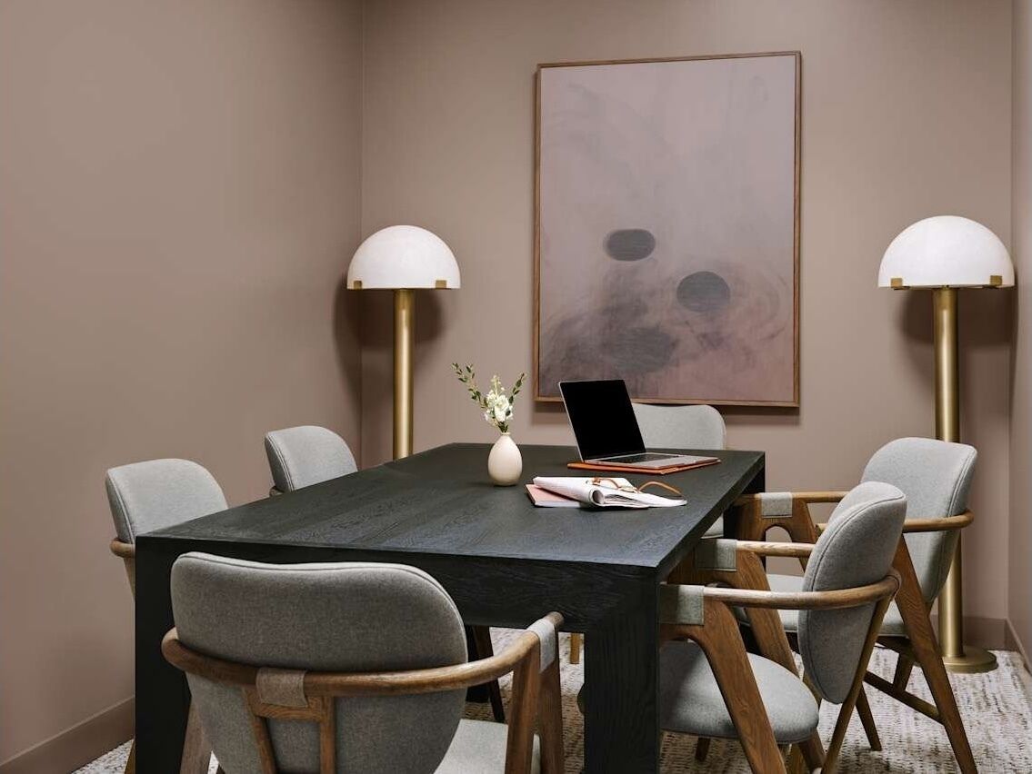 A conference room with a dark wooden table, grey upholstered chairs, a laptop, and two floor lamps.