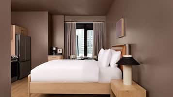 Habitación de hotel con cama, mesita de noche, lámpara y vista a un paisaje urbano a través del ventanal.
