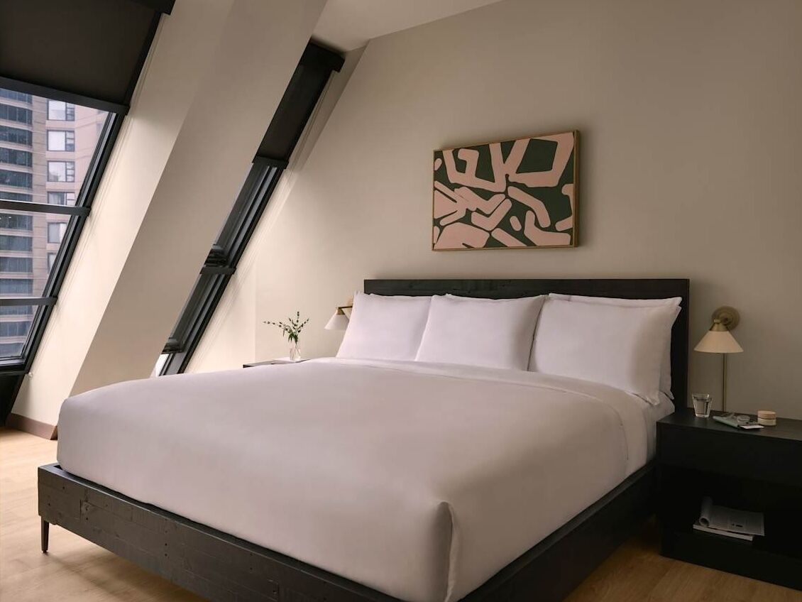 A modern bedroom with a large bed, a nightstand, a wall-mounted abstract art piece, and a window with a view of buildings.