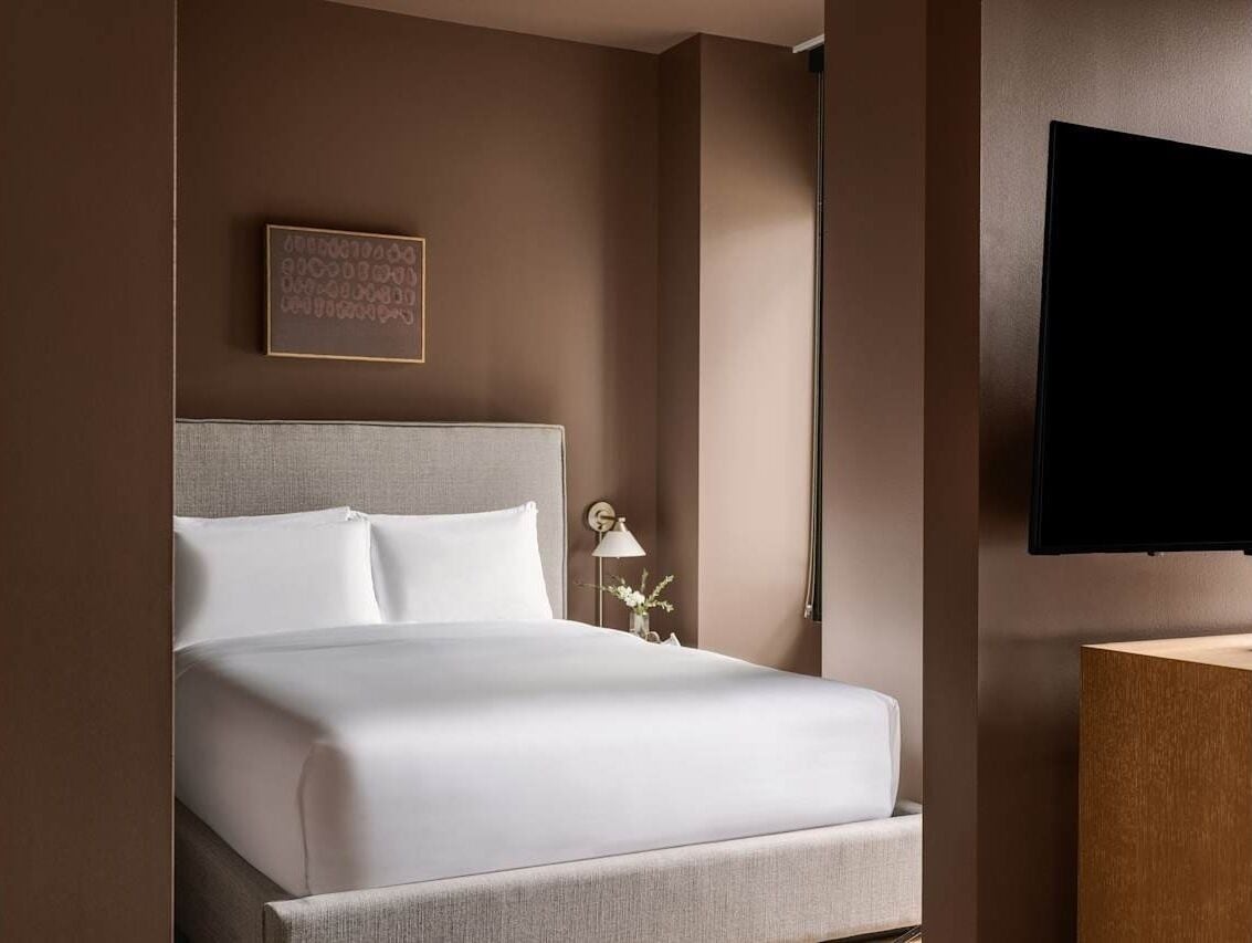 Une chambre d’hôtel avec un lit, un téléviseur fixé au mur, une table de chevet avec une lampe et un tableau encadré accroché au mur.