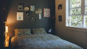 A bedroom with a bed, a bedside lamp, a window, and various wall art. - Artist’s EcoRetreat in the heart of Ballard (Seattle)