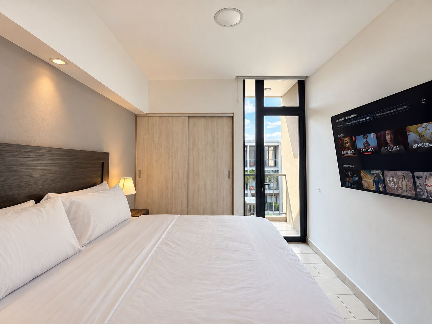 A modern hotel room with a large bed, a nightstand, a lamp, a sliding door to a balcony, and a wall-mounted TV displaying a selection of movies and shows.