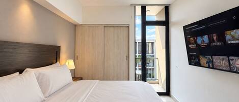 A modern hotel room with a large bed, a nightstand, a lamp, a sliding door to a balcony, and a wall-mounted TV displaying a selection of movies and shows.