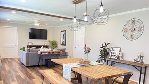 Un salon moderne avec une table à manger en bois, des bancs et un canapé. On y trouve des luminaires suspendus, une horloge murale et un téléviseur à écran plat.