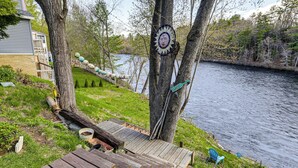 Une terrasse en bois menant à la rive d’une rivière, avec des chaises et un objet décoratif accroché à un arbre.