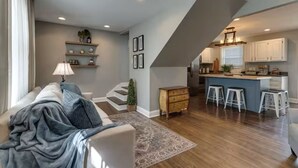 Una sala de estar moderna con un sofá, una mesa de centro, una lámpara, una escalera y una zona de cocina con taburetes de bar.