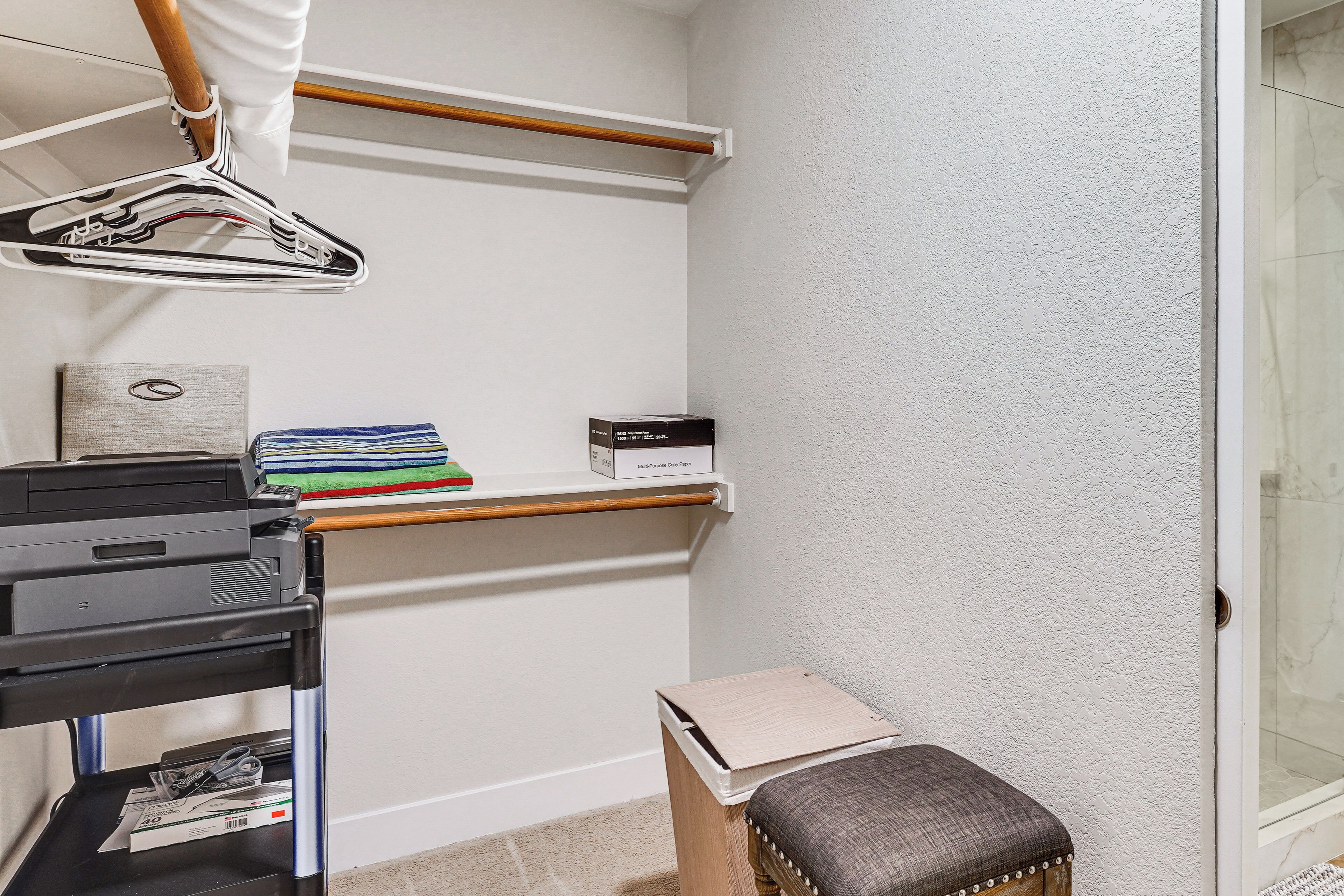 A corner of a room with a printer, a small table with a cushion, a hanging garment rack, and a shelf with folded clothes and a box.