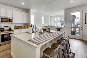 A modern kitchen with white cabinetry, stainless steel appliances, a central island with bar stools, and a view through the door. - Pets Welcome! Thornton Extended Stay w/ Balcony (Thornton)