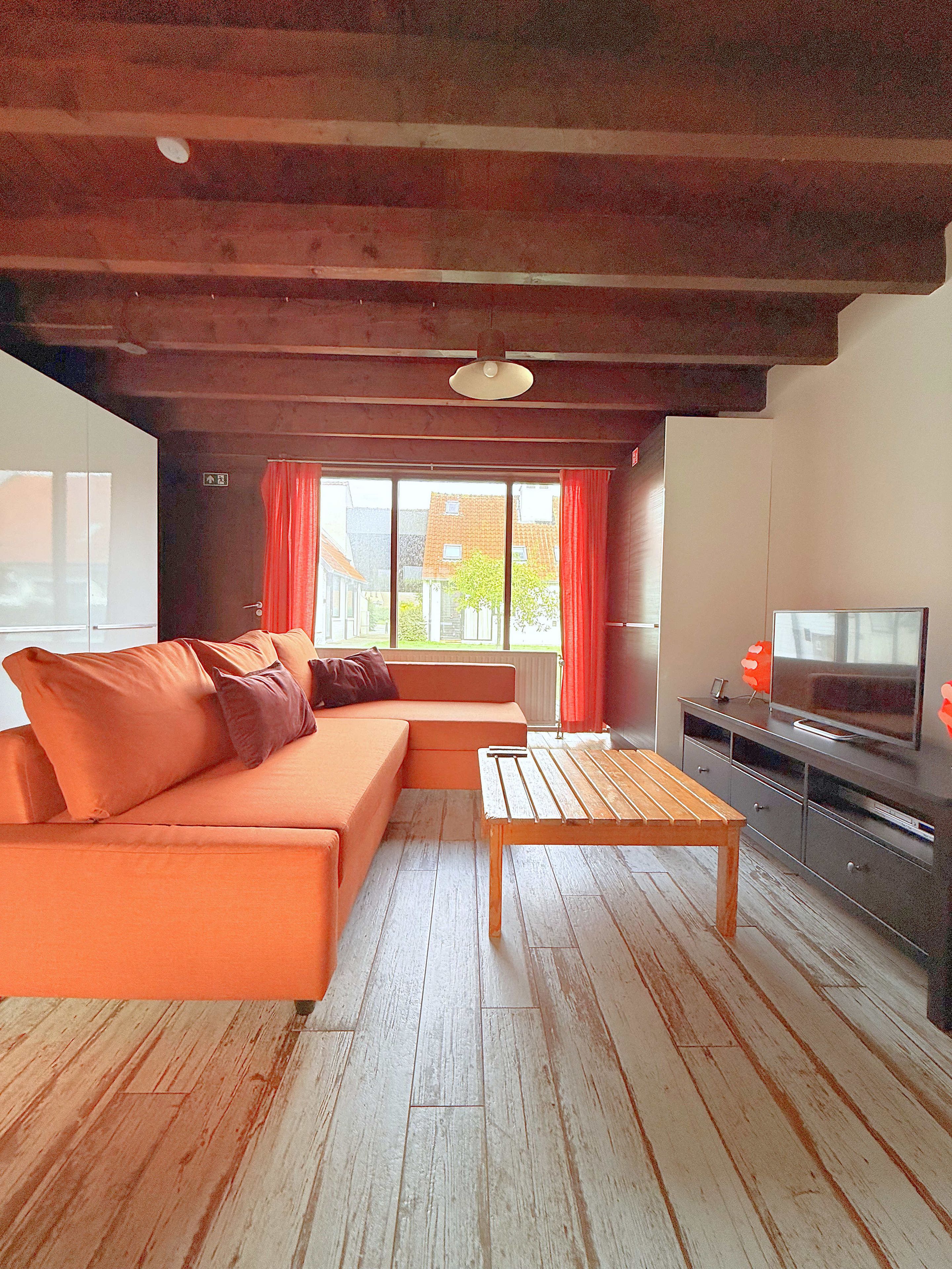 Un salon moderne avec un plafond en bois, un grand canapé orange, une table basse en bois et un téléviseur posé sur un meuble.