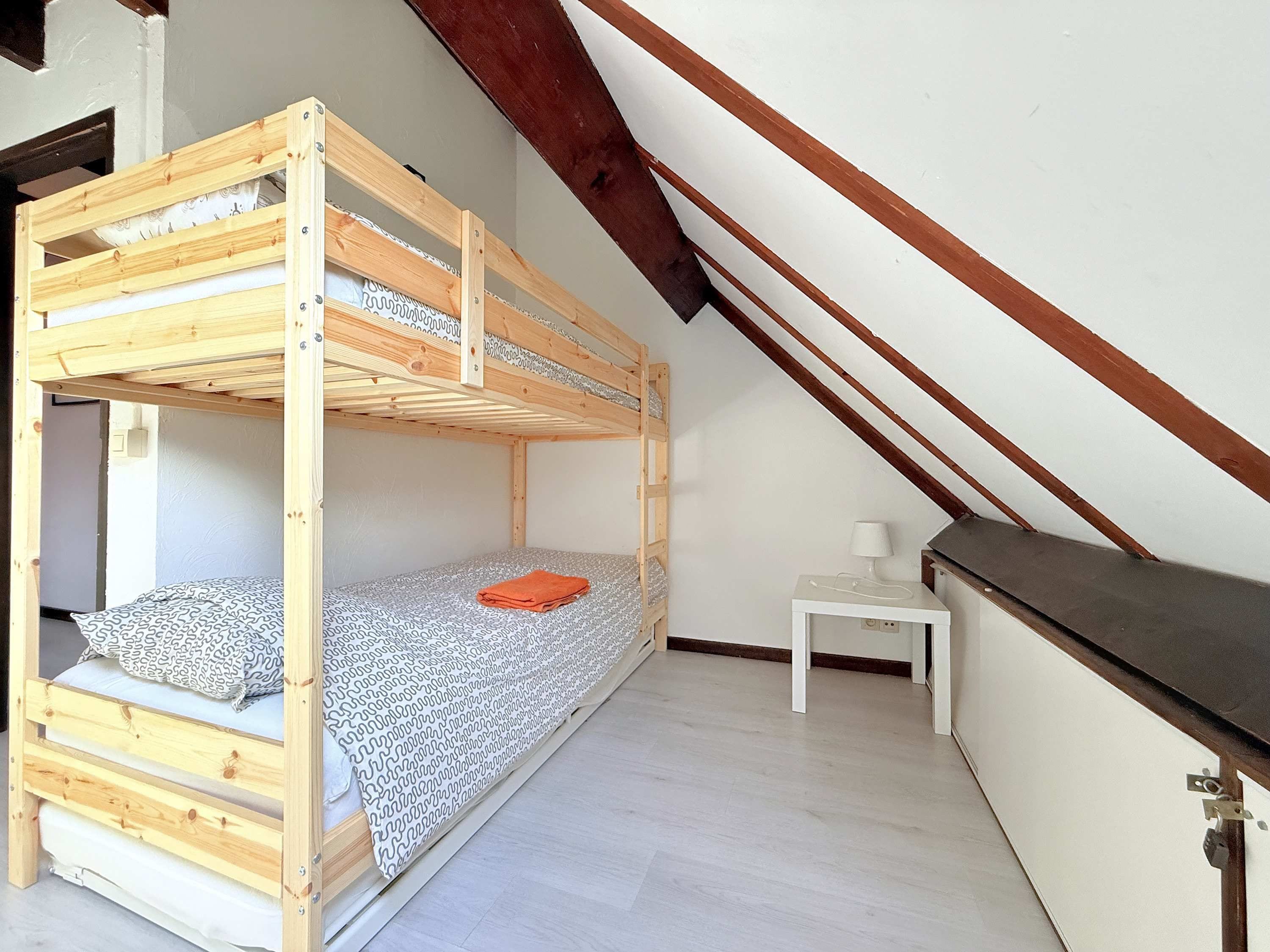 Un lit superposé dans une pièce avec un plafond en pente, une petite table de chevet avec une lampe et un mur blanc.