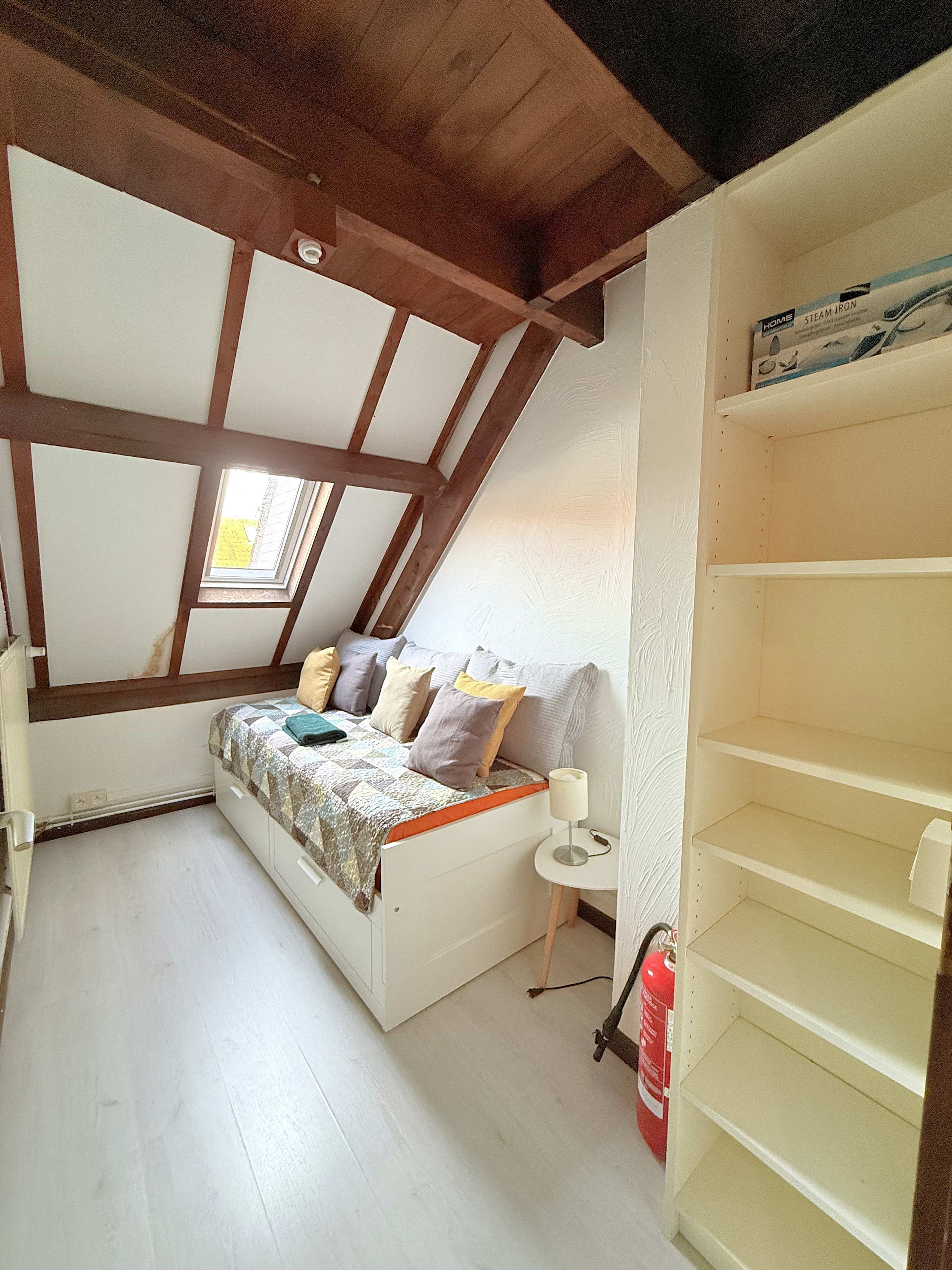 Une chambre avec un plafond en pente, un lit recouvert d’une courtepointe, une table de chevet avec une lampe et un extincteur.