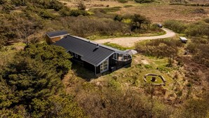Ein modernes Haus mit einer großen Terrasse, eingebettet in eine natürliche Landschaft mit Hügeln und spärlicher Vegetation.