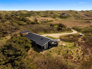Ein modernes Haus mit einer großen Terrasse, eingebettet in eine natürliche Landschaft mit Hügeln und spärlicher Vegetation.