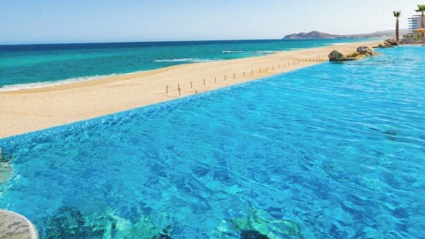 A clear blue pool with a sandy beach and a building with palm trees. - Luxury Cabo Escapes Suites, Villas & Elevated Experiences by Chez Tandrie (San José del Cabo)