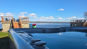 Une piscine entourée d’une terrasse en pierre, une autre terrasse en bois avec des chaises, et une vue sur l’eau.