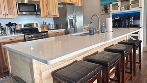 Une cuisine moderne avec des meubles en bois, des appareils électroménagers en acier inoxydable, un grand îlot central doté de tabourets, et une vue sur l’océan par une fenêtre.