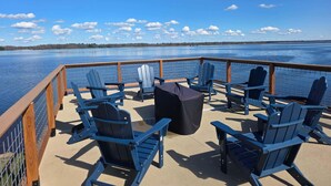 Une terrasse avec des chaises Adirondack bleues et une table couverte, donnant sur un lac paisible sous un ciel dégagé.