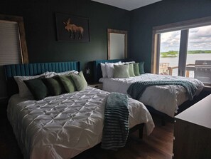 A hotel room with two beds, a moose artwork, a view of the water, and a wooden deck outside.