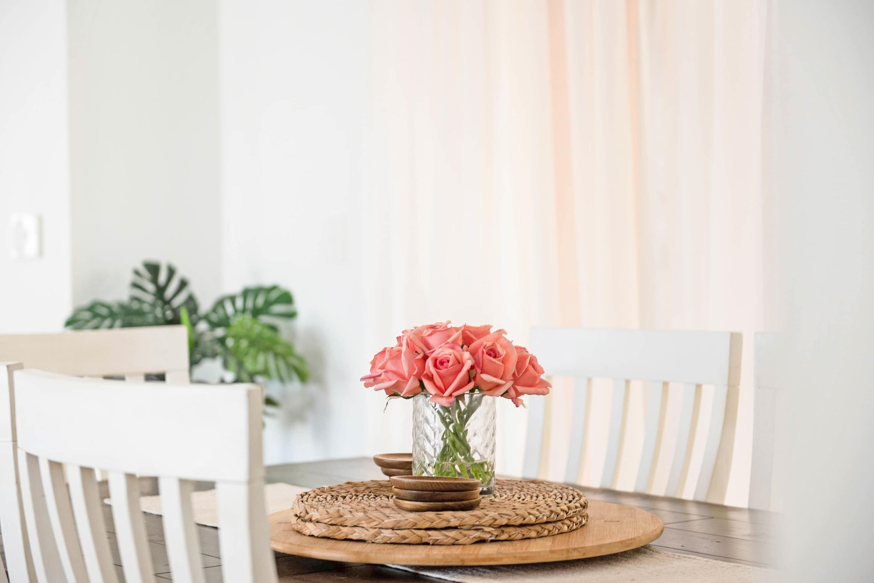 A dining table with a vase of pink roses, a woven placemat, and a white chair.
