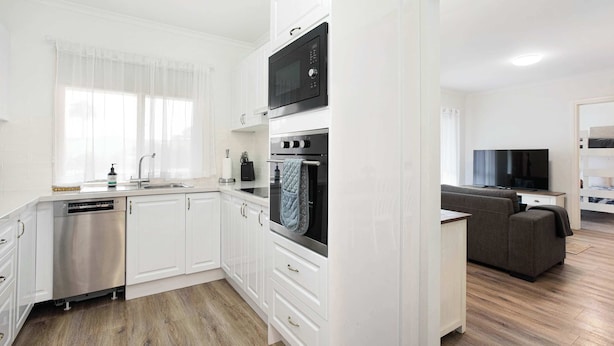 Una cocina moderna con armarios blancos, lavavajillas, microondas y horno. En la sala de estar, hay un sofá gris y un televisor de pantalla plana.