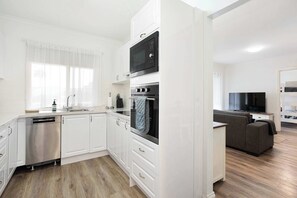 A modern kitchen with white cabinetry, a dishwasher, a microwave, and an oven. There is a grey sofa and a flat-screen TV in the living area. - Kingsford Cottage, Walk to Beach, Dog Friendly (Victor Harbor)
