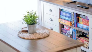 Una habitación con una mesa de madera, una planta en maceta, una lámpara y una estantería con juegos de mesa.