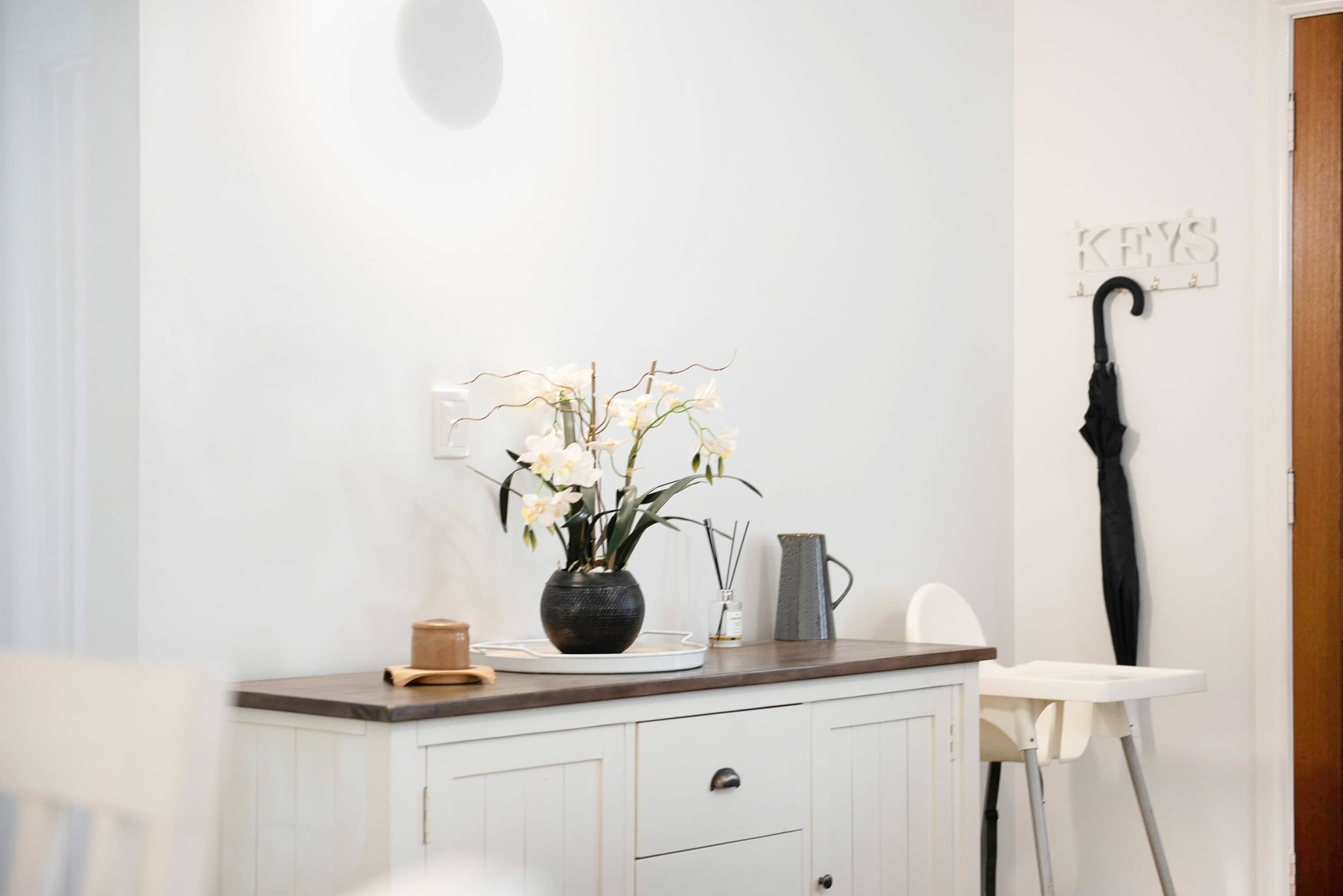 A white wall with a black vase of white flowers, a white chair, a white cabinet, and a black umbrella hanging on the wall.