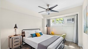 Un dormitorio con cama, mesita de noche, lámpara, ventilador de techo, ventana y aire acondicionado.