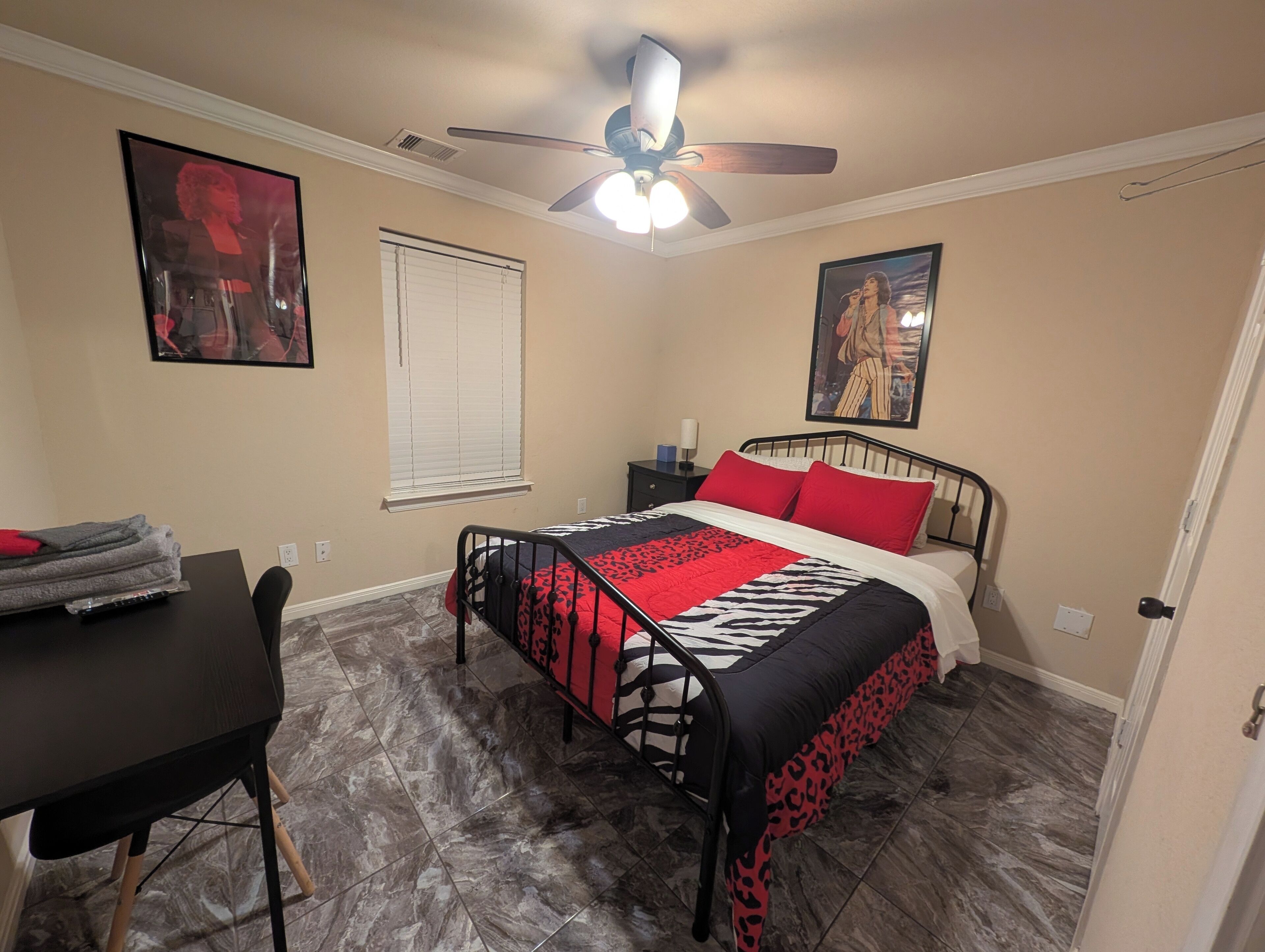 Een slaapkamer met een bed, een bureau, een stoel, een plafondventilator en twee ingelijste foto’s aan de muur.
