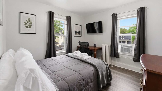 A bedroom with a bed, a desk, a chair, a TV, and large windows with curtains. - The Grandstands Apt 2 - All Star Baseball Rentals (Oneonta)