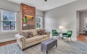 Uma sala de estar com um sofá bege, uma poltrona verde, uma mesinha com um abajur e uma lareira com parede de tijolos.