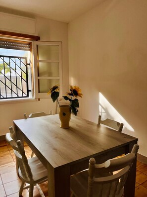 A dining area with a wooden table and chairs, a window with blinds, and a potted plant with sunflowers. - Your corner of paradise just a stone's throw from the sea, among dunes and the scent of Salento 🌾 (Puglia)