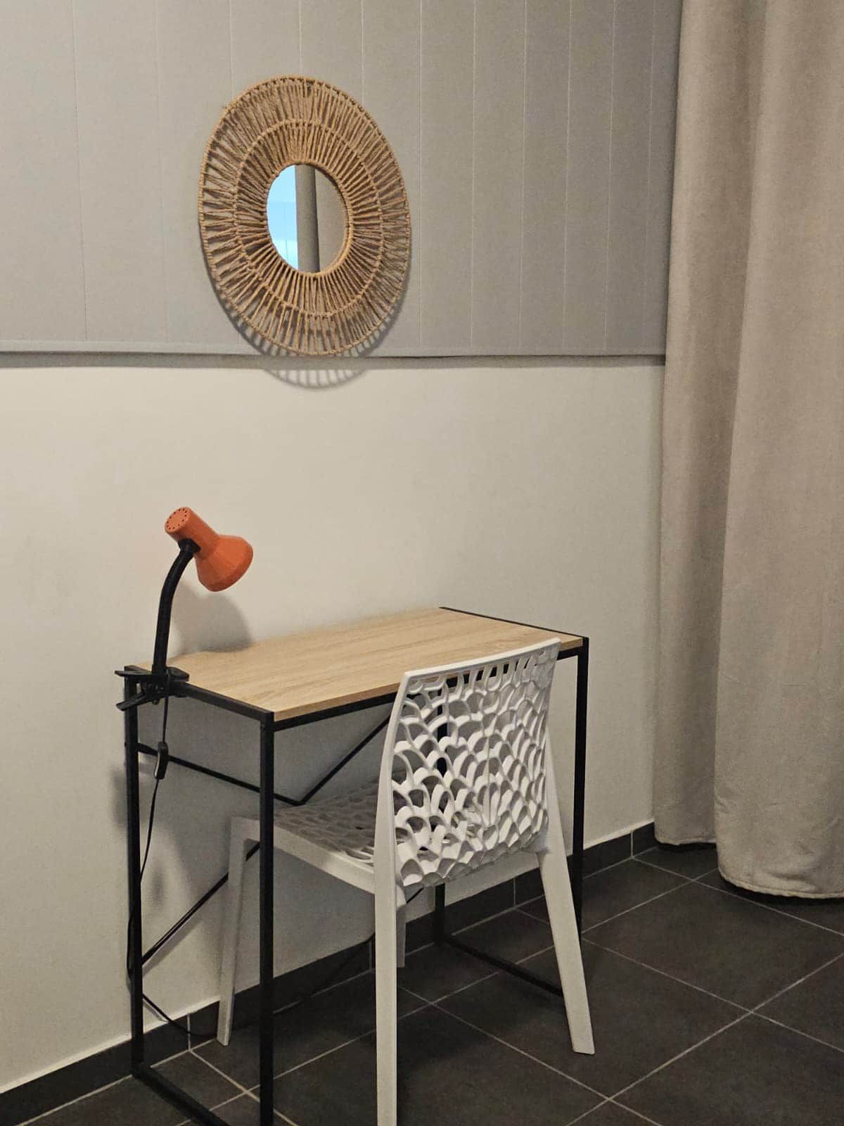 Une petite pièce moderne avec une table en bois et une chaise blanche, un miroir rond en osier accroché au mur et un rideau à la fenêtre.