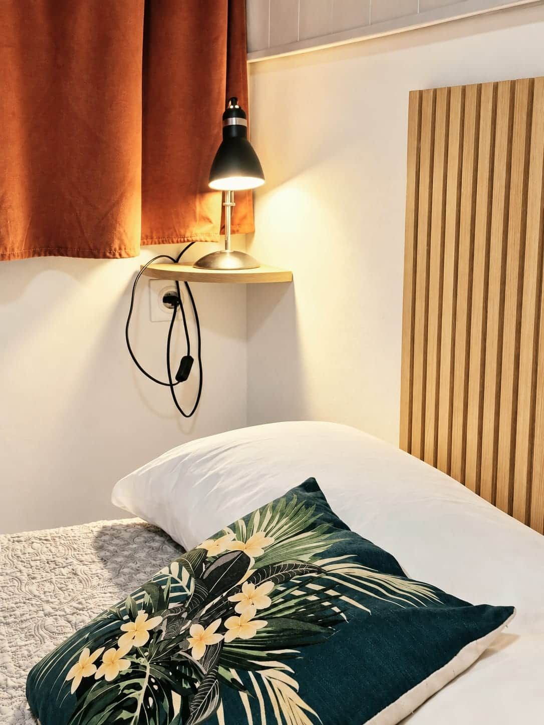 A bed with a white pillow and a green pillow with a tropical design, a bedside lamp, and a wooden headboard.