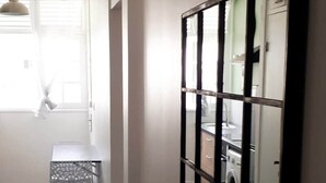 Ein Zimmer mit Fliesenboden, einem Metallstuhl, einem Fenster mit Jalousien und einem wandmontierten Spiegel.