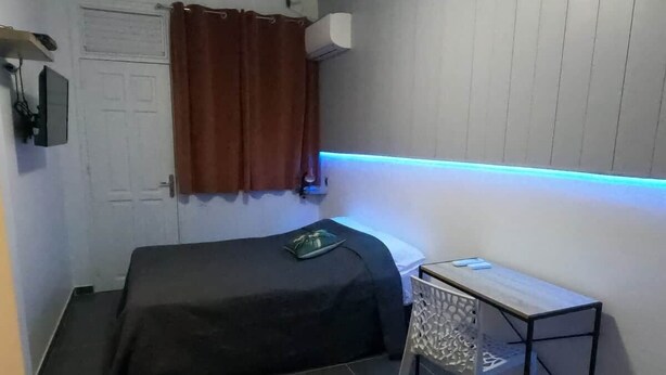 Ein kleines Zimmer mit einem Bett, einem Tisch, einem Stuhl, einem Vorhang und einer Klimaanlage.