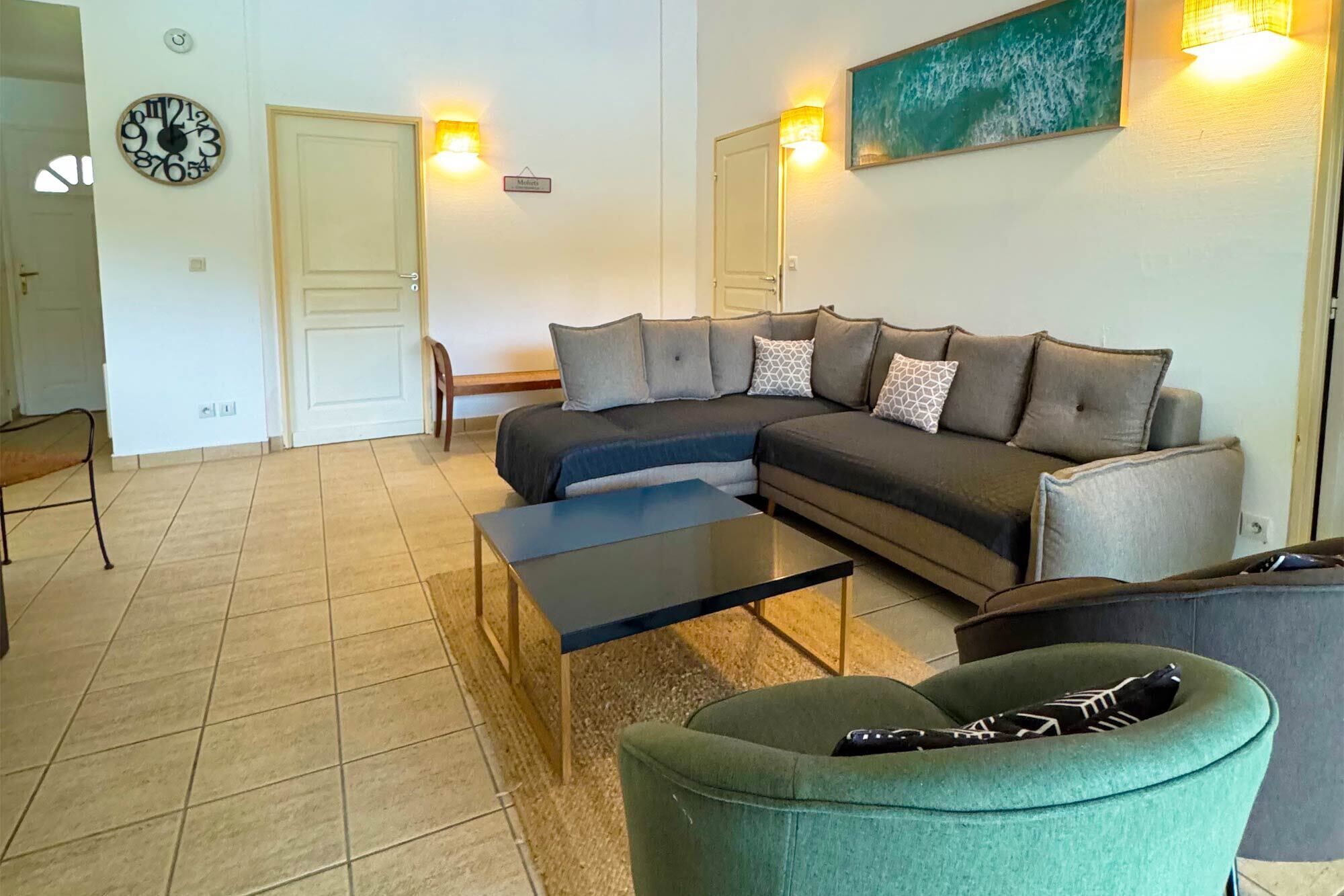 Ein Wohnzimmer mit einem Ecksofa, einem Couchtisch, einem grünen Sessel, einer Wanduhr und zwei Wandleuchten.