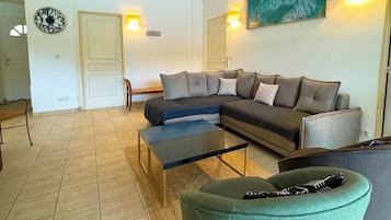 Ein Wohnzimmer mit einem Ecksofa, einem Couchtisch, einem grünen Sessel, einer Wanduhr und zwei Wandleuchten.